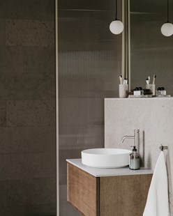 Baderomsinnredning i tre med hvit bolleservant, kalksteinsvegg og dusjvegg i rillet glass.