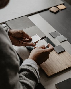 Hender som vurderer material- og fargeprøver til baderom, med fliser, metallprøver og treverk lagt utover et bord.